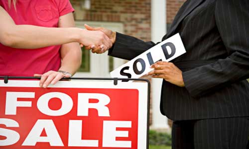 People shake hands after a house sale. Person on left holds a red "For Sale" sign. Person on the right holds a white "Sold" sign.
